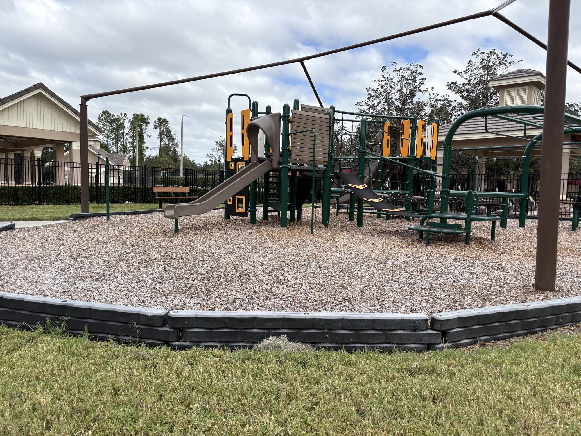 The view from the bench: the playground on gravel under a metal shade frame, with a pavilion behind.