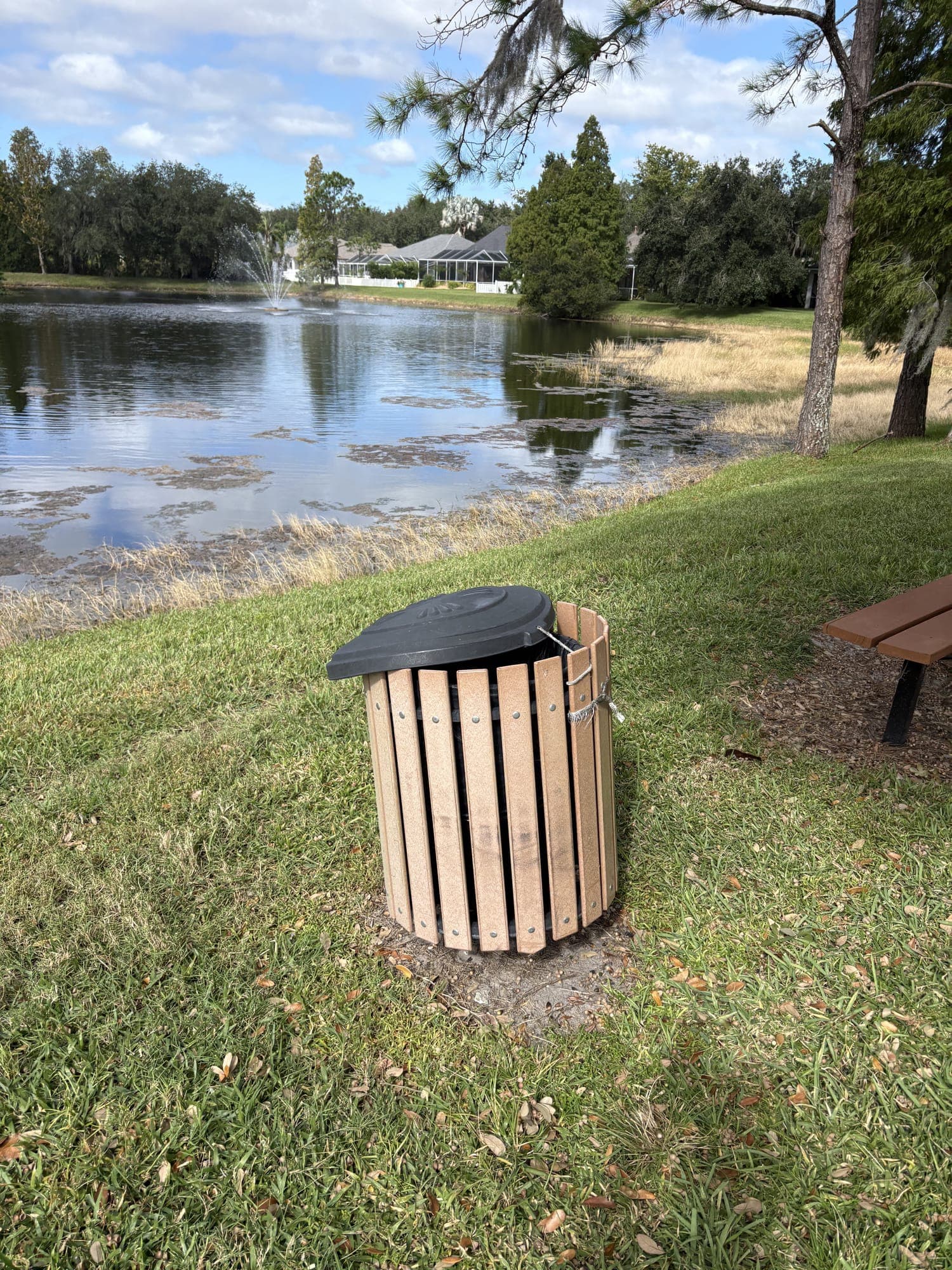 The wooden-slat trash can on the grass right beside the bench, with the pond beyond.