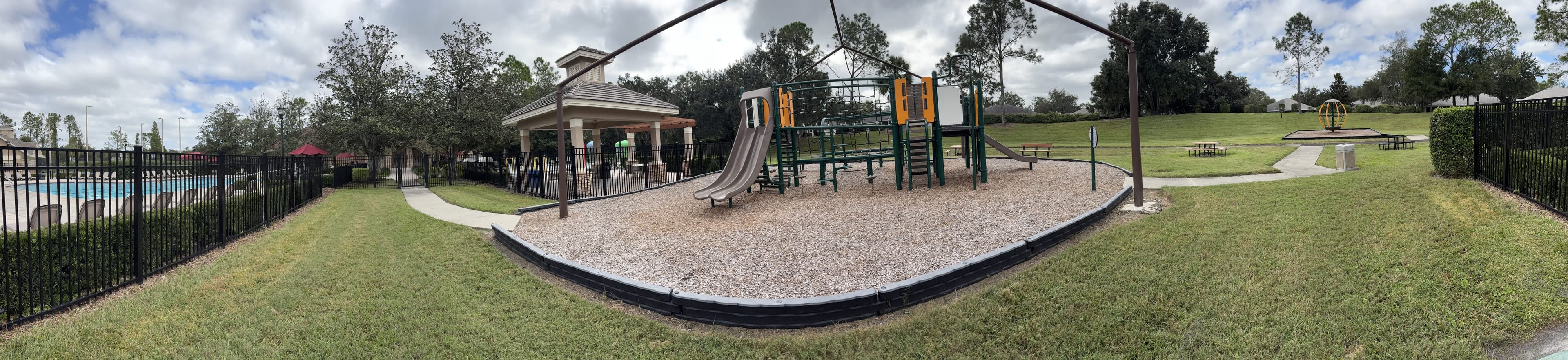 Panorama from the bench: the pool fence on the left, a pavilion and the playground in the middle, a wide lawn on the right.