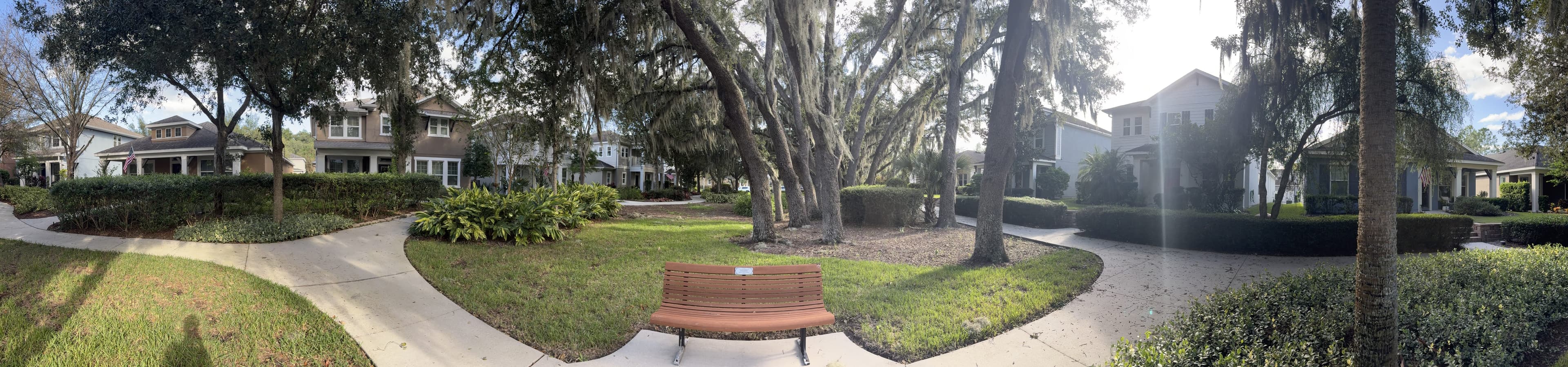 Panorama from the bench: paths curving around a round lawn with oaks, the other bench in the middle, and houses all around.