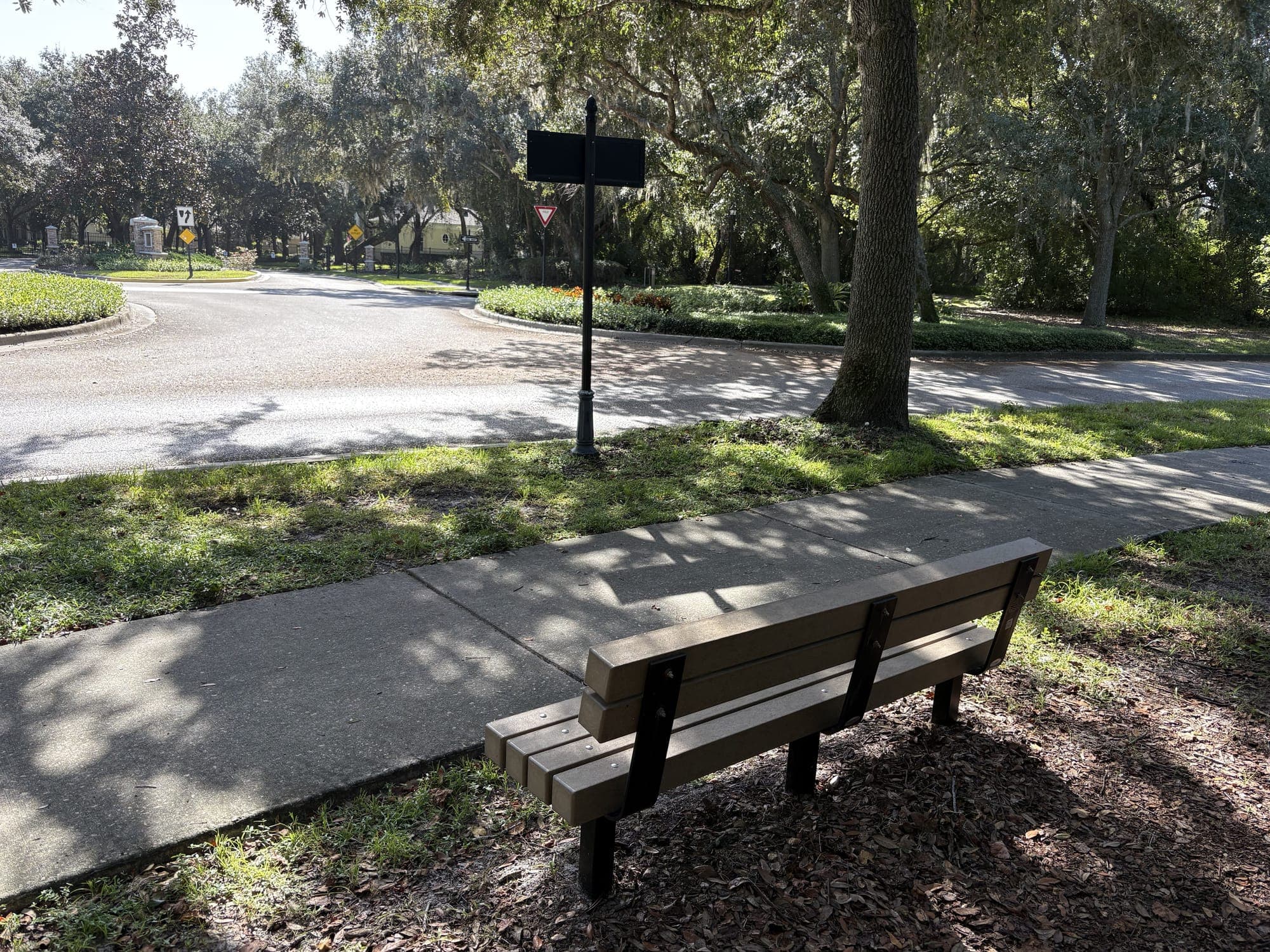 The bench beside the sidewalk with the roundabout, a sign post, and oak trees across the road.