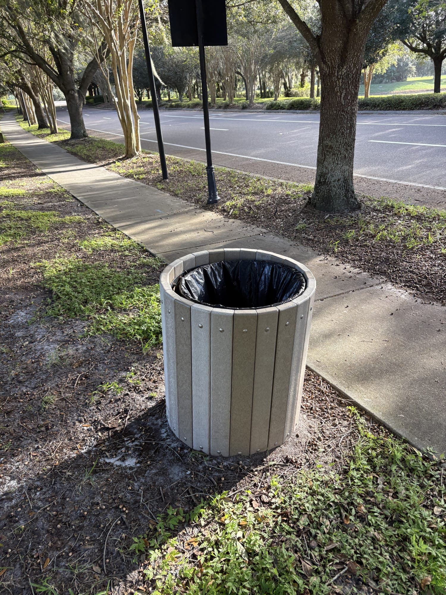 The gray trash can beside the bench, on the grass by the sidewalk.
