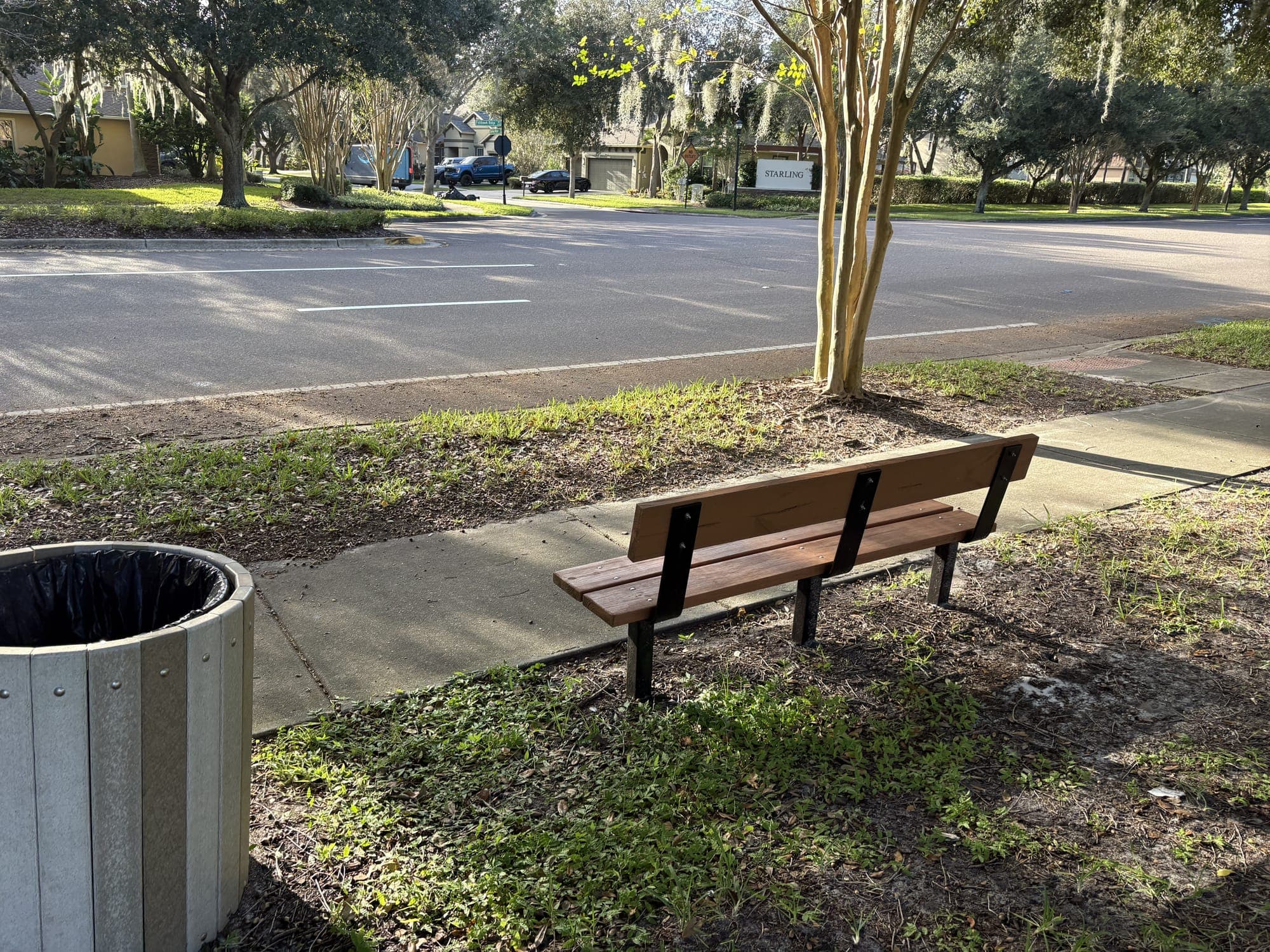 The bench along the sidewalk with the trash can next to it and the road ahead.