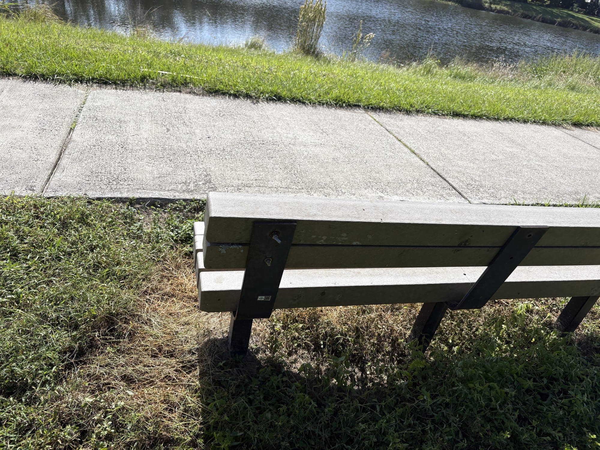 The end of the bench beside the sidewalk, with the pond just beyond the grass.