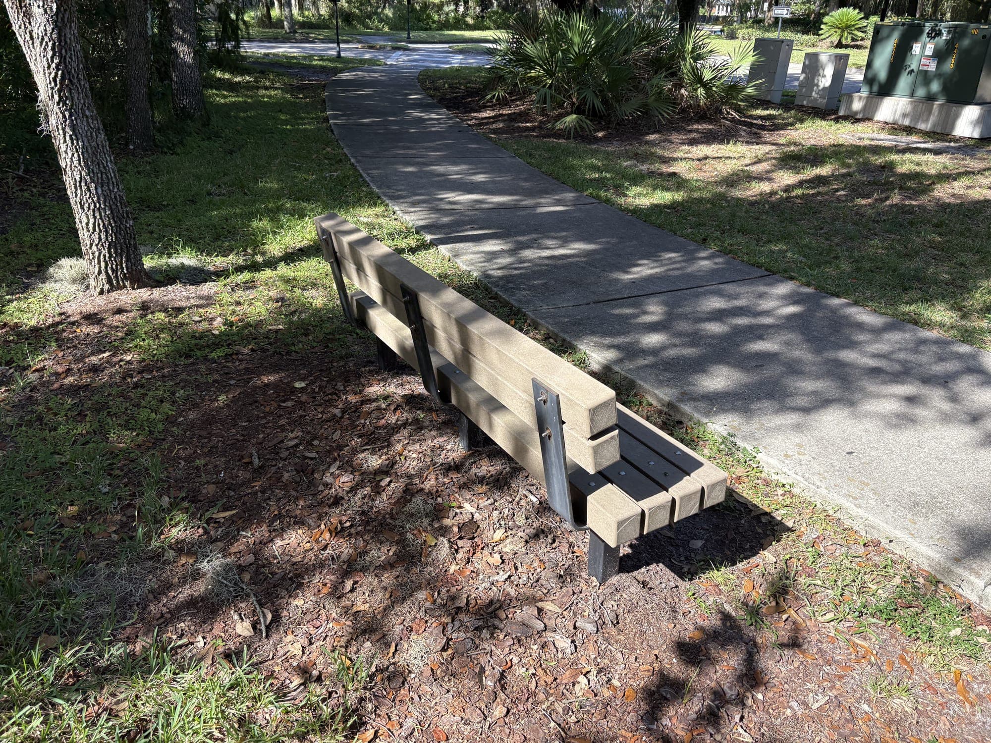 The bench from behind, beside a sidewalk under oaks.