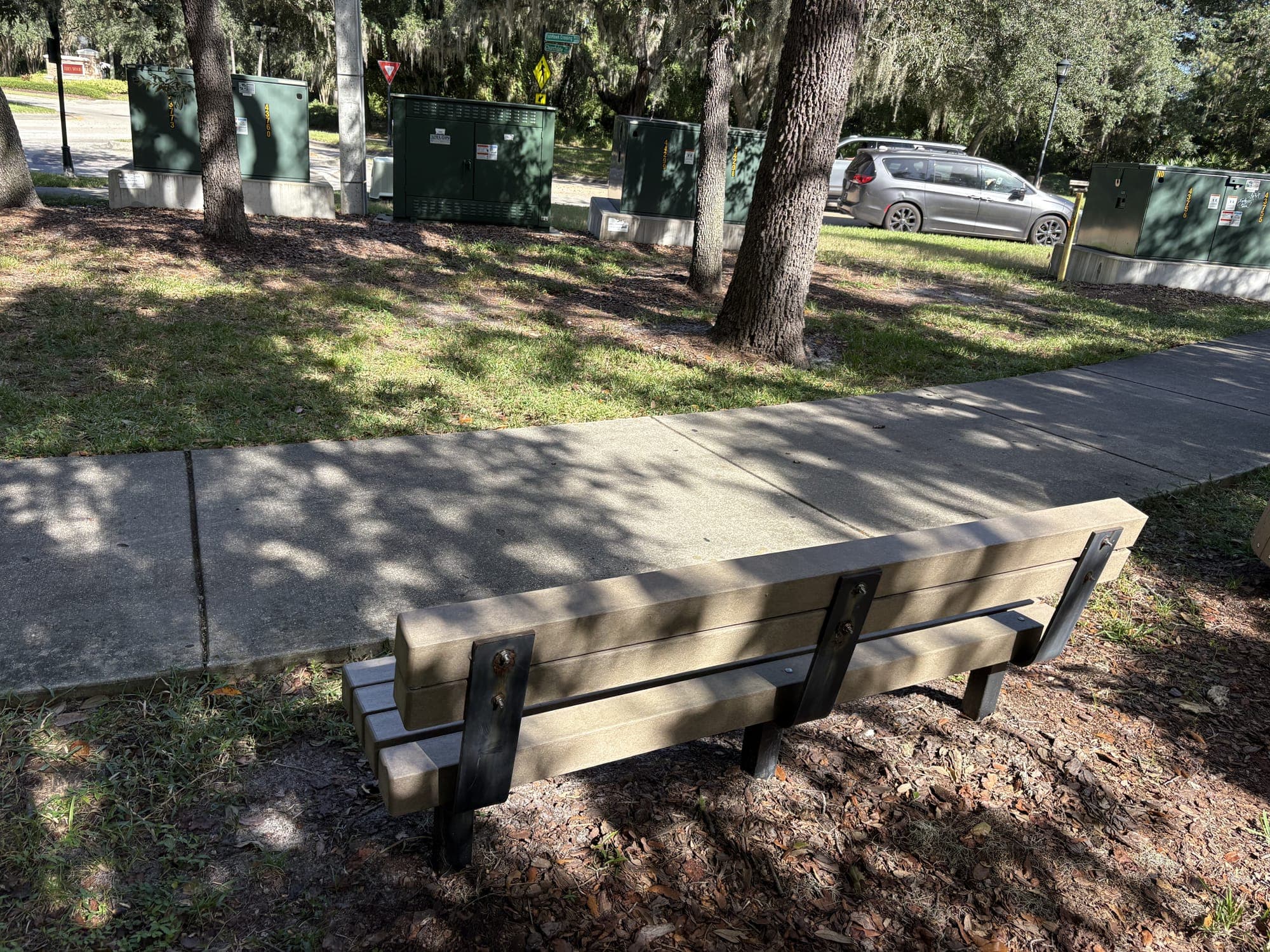 The bench facing a lawn with green utility boxes and a parked car beyond the trees.