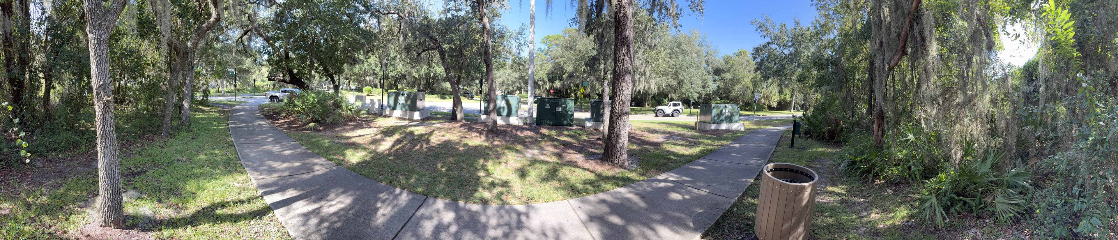 Panorama around the bench: oaks, the curving path, utility boxes and parked cars, and a trash can on the right.