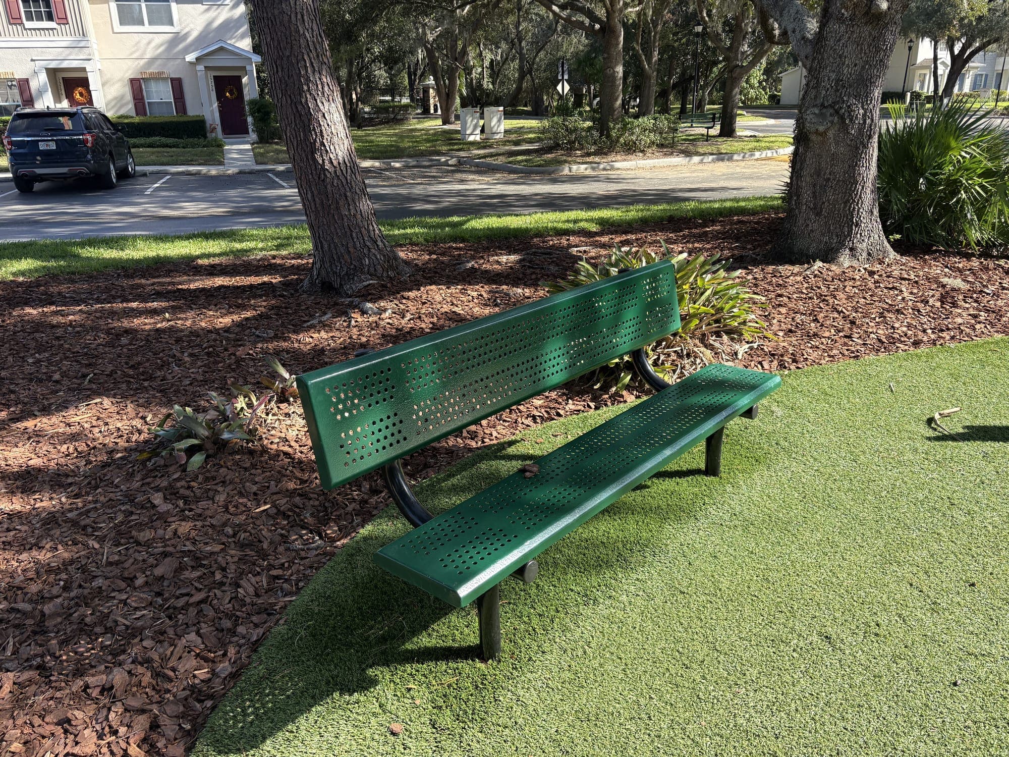The green bench on turf, with mulch, oaks and the street with parked cars behind it.