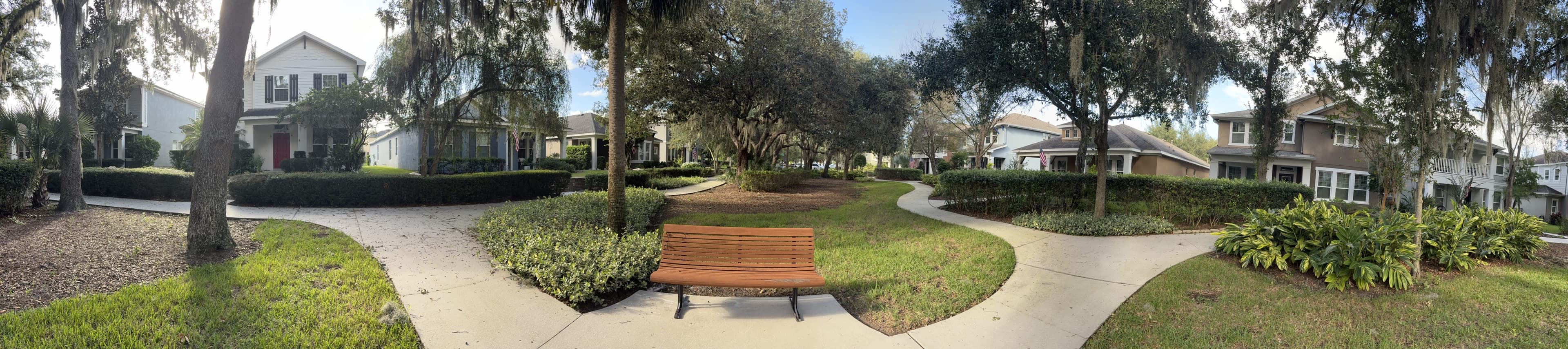Panorama from the bench: paths and hedges around a round lawn, the other bench in the middle, houses beyond.