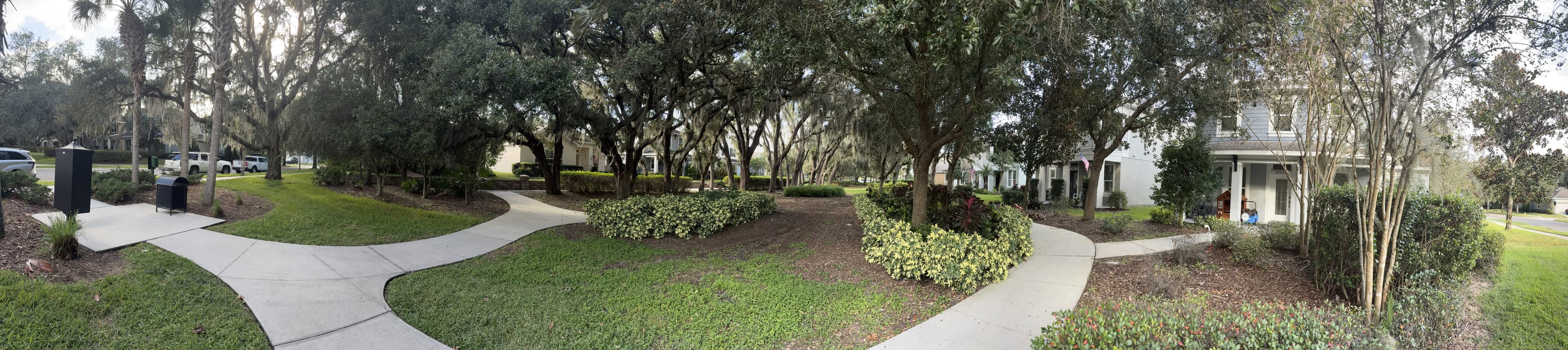 Panorama from the bench: sidewalks curving through lawns and flower beds under oaks, houses on the right.